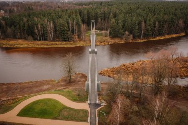 Valmiera, Latvia - November 14, 2024 - Aerial view of a suspension bridge over a river, leading into a dense forest, with a winding pathway and bare trees in autumn tones. clipart