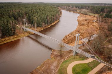 Valmiera, Letonya - 14 Kasım 2024 - Çağdaş bir asma köprünün dolambaçlı bir nehir üzerindeki havadan görünüşü, yoğun orman ve sonbahar manzarasıyla çevrili, sakin bir doğal manzara yaratıyor..
