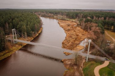 Valmiera, Letonya - 14 Kasım 2024 - Orman ve sonbahar manzarası ile çevrili sakin bir nehri geçen bir asma köprünün havadan görünüşü.