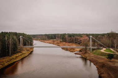 Valmiera, Letonya - 14 Kasım 2024 - Çevresindeki sonbahar ağaçları, tarlaları ve bulutlu gökyüzü ile sakin bir doğal manzara oluşturan sakin bir nehri geçen bir asma köprünün havadan görünüşü.