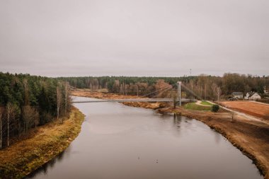 Valmiera, Letonya - 14 Kasım 2024 - Çevresindeki sonbahar ağaçları, tarlaları ve bulutlu gökyüzü ile sakin bir doğal manzara oluşturan sakin bir nehri geçen bir asma köprünün havadan görünüşü.