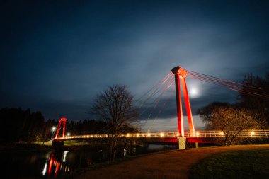 Valmiera, Letonya - 15 Kasım 2024 - Vansu eğilir. Geceleri parlak kablolar, ay ışığı ve sudaki yansımalarla aydınlatılan kırmızı köprü huzurlu ve modern bir gece manzarası yaratıyor..