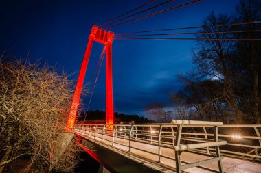 Valmiera, Letonya - 15 Kasım 2024 - Vansu eğilir. Koyu mavi gökyüzüne karşı koyu kırmızı ışık saçan modern bir yaya köprüsü, parlak tasarımı ve mimari çizgileri vurguluyor..