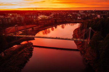 Valmiera, Letonya - 15 Kasım 2024 - Vansu eğilir. Nehir üzerindeki bir köprünün havadan görünüşü canlı turuncu günbatımını yansıtıyor, arka planda şehir ışıkları ve binalar görünüyor..