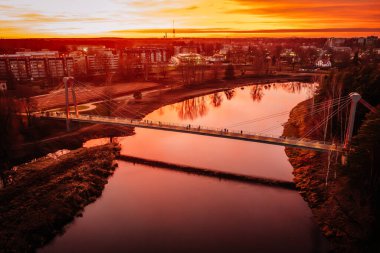 Valmiera, Letonya - 15 Kasım 2024 - Vansu eğilir. Nehir üzerindeki bir köprünün havadan görünüşü canlı turuncu günbatımını yansıtıyor, arka planda şehir ışıkları ve binalar görünüyor..