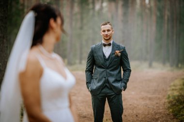 Koknese, Letonya - 26 Eylül 2023 - Özel dikim bir takım elbise içindeki damat, ormanın içinde kendine güvenle duruyor, yumuşak odak noktasında geline bakıyor, romantik bir açık hava düğün atmosferi yaratıyor..