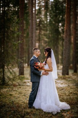 Koknese, Letonya - 26 Eylül 2023 - Bir gelin ve damat ormanda samimi bir anı paylaşıyor, birbirlerine sarılıp sevgiyle bakıyorlar. Gelin bir buket tutuyor ve güneş ışığı ağaçların arasından süzülüyor..