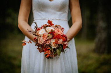 Koknese, Letonya - 26 Eylül 2023 - Sonbahar temalı bir buket gül, yıldız çiçeği ve okaliptüs yapraklı, kırmızı, şeftali ve krema renginde beyaz bir gelinlik içinde gelin.