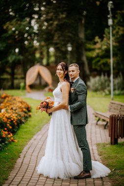 Gelin ve damat rengarenk çiçeklerle ve yeşilliklerle bir bahçe yolunda duruyorlar, düğün günlerinde kucaklaşıyorlar ve uzaklara bakıyorlar..