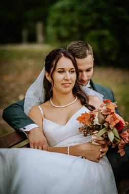 Koknese, Latvia - September 26, 2023 - Bride and groom share an intimate moment on a bench, with the bride holding a bouquet and the groom embracing her from behind, capturing their love. clipart