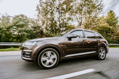 Koknese, Letonya - 26 Eylül 2023 - Düğün için çiçeklerle süslenmiş, arka planda bulanık ağaçlarla dolu manzaralı bir yolda giden koyu renkli bir SUV.