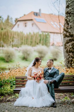 Gelin ve damat parkta bir bankta oturuyor, arka planda güzel bir bahçe ve kırsal evle birbirlerine gülümsüyorlar, romantik bir düğün sahnesi yaratıyorlar..