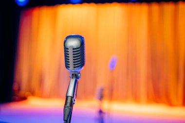 Arka planda bulanık altın bir perde ve mavi sahne ışıkları olan bir tribünde gümüş bir mikrofon. Performans temaları için mükemmel.