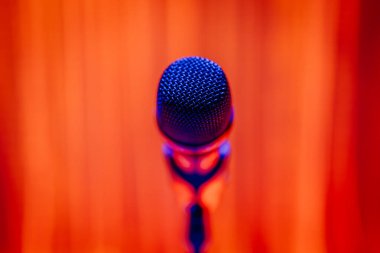 Canlı turuncu ve mavi ışıklandırmalı bir standda mikrofonun yakın çekimi. Müzik, performans ve topluluk konuşması için mükemmel bir tema..