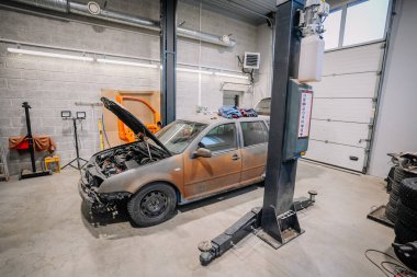 Valmiera, Letonya - 18 Kasım 2024 - Bir garaj tamirhanesinde kaputu açık bir araba, etrafında aletler, asansör ve duvarda turuncu bir araba kapısı var..