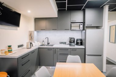 Valmiera, Letonya - 18 Kasım 2024 - Siyah dolaplı şık bir ofis mutfağı, bir tezgah, bir lavabo, bir mikrodalga fırın, bir buzdolabı, bir kahve makinesi ve duvara monte edilmiş bir televizyon.
