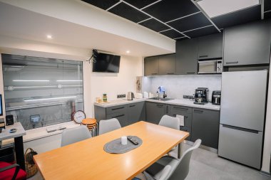 Valmiera, Letonya - 18 Kasım 2024 - Siyah dolaplı modern bir ofis mutfağı, ahşap bir masa, gri sandalyeler, buzdolabı, lavabo, cihazlar ve duvara monte edilmiş bir televizyon..