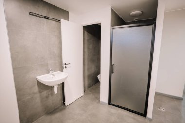 A modern bathroom with gray tiled walls, a sink, a frosted glass shower enclosure, and a partially open door leading to a toilet area. clipart