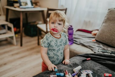 Yüzünde dağınık ruj izi olan, kozmetiklerle çevrili, neşeli ve meraklı bir ifadeyle rahat bir odada duran bir çocuk..