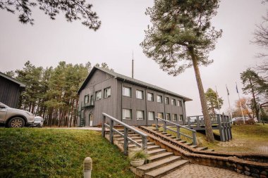 Valmiera, Letonya - 19 Kasım 2024 - Merdivenleri, çam ağaçları, çimenler ve bulutlu bir gökyüzünün altında park etmiş arabalarla çevrili iki katlı, gri ahşap bir bina.