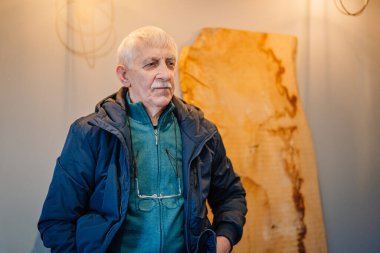 Valmiera, Latvia - November 19, 2024 - An elderly man wearing a jacket and sweater, standing indoors with a thoughtful expression, rustic wooden decor in the background.