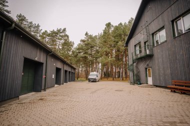 Valmiera, Letonya - 19 Kasım 2024 - Gri ahşap binalar, park edilmiş bir minibüs, spiral merdivenler ve etrafındaki çam ağaçları ile kaplı bir avlu.