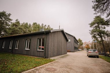 Valmiera, Letonya - 19 Kasım 2024 - Asfaltlı garaj yolu, park edilmiş araba, arka planda ağaçlar ve bulutlu bir günde görünen bayrakları olan modern ahşap bina.