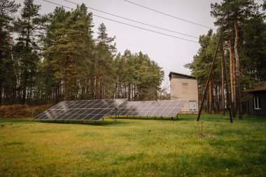 Valmiera, Letonya - 19 Kasım 2024 - Bir ormanın yakınındaki çimenli bir alanda güneş panelleri, elektrik direkleriyle ve bulutlu bir gökyüzünün altında küçük bir tuğla bina.