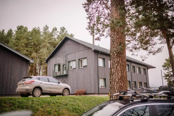 Valmiera, Letonya - 19 Kasım 2024 - Çam ağaçları ve bulutlu gökyüzünün altında yeşil çimlerle çevrili arabaları olan modern gri ahşap bir bina.