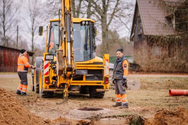 Valmiera, Latvia - November 19, 2024 - Two workers in high-visibility gear next to a yellow excavator and an open manhole at a construction site in a rural setting. clipart