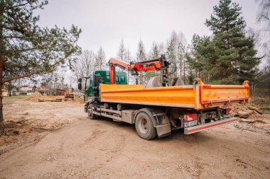 Valmiera, Letonya - 19 Kasım 2024 - Hidrolik vinç ve turuncu yataklı, ağaçlar, kum ve makinelerle çevrili bir Volvo kamyonu olan bir inşaat alanı.