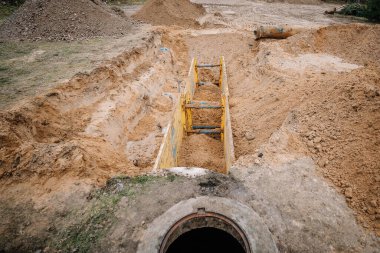 Valmiera, Letonya - 19 Kasım 2024 - Kum yığınları ve büyük bir boru ile çevrili sarı destekli bir inşaat sahasında açık siper. Yararlı iş için kazı.
