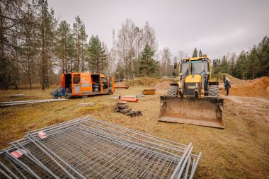 Valmiera, Letonya - 19 Kasım 2024 - Sarı bir kazıcı, turuncu bir minibüs, metal çit, boru ve ormanlık alanda çalışan bir işçi.