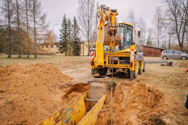 Valmiera, Letonya - 19 Kasım 2024 - Kum ve ağaçlarla çevrili bir inşaat sahasında zincir kancalı bir kazıcı çalışıyor.