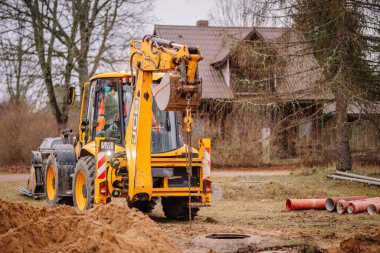 Valmiera, Letonya - 19 Kasım 2024 - Sarı bir kepçe yükleyici ön planda borular ve toprak taşıma ekipmanları ve arka planda ağaçlar olan bir inşaat alanında kazı yapıyor.