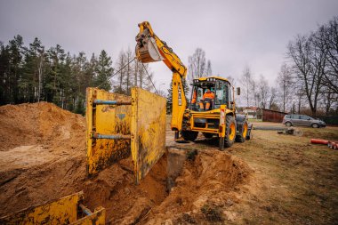 Valmiera, Letonya - 19 Kasım 2024 - Bir kepçe yükleyici, arka planda toprak yığınları ve ağaçların bulunduğu bir inşaat alanında sarı siper kalkanını kaldırıyor..