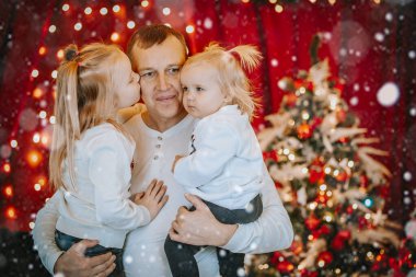 Bir baba Noel kutlamasında iki kızını kucağına alır. Bir kızı onu yanağından öperken diğeri ona sevgiyle bakar..