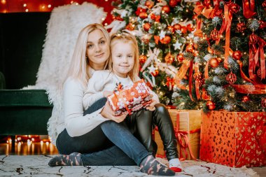 Süsler ve tatil ışıklarıyla çevrili kırmızı bir hediye kutusuyla süslü bir Noel ağacının yanında oturan bir anne ve kız..
