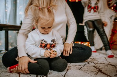 Küçük bir çocuk festival ortamında yerde oturur, küçük bir oyuncakla oynar, tatil temalı bir kazak giyer, yakınlarda bir aile üyesiyle birlikte..