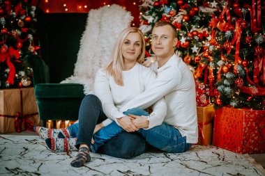 Güzel dekore edilmiş bir Noel ağacının yanında oturan bir çift. Etrafı şenlikli süsler, hediyeler ve hoş bir tatil dekoruyla çevrili..