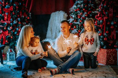 Valmiera, Letonya - 23 Kasım 2024 - Noel 'i havai fişeklerle kutlayan neşeli bir aile, Noel süsleri, Noel ağaçları ve kar taneleri efektleri ile çevrili, sıcak bir tatil ortamı yaratıyor..