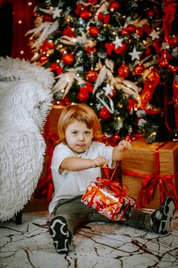 Genç bir çocuk süslenmiş bir Noel ağacının yanında yerde oturuyor. Parlak bir Noel dekoruyla çevrili kırmızı bir hediye kutusunun kurdelesini çözüyor..