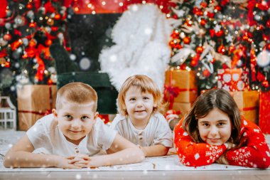 Üç gülümseyen çocuk, parlayan ışıkları ve paketlenmiş hediyeleriyle bir Noel ağacının yanında uzanmış bayram sezonunun tadını çıkarıyorlar..