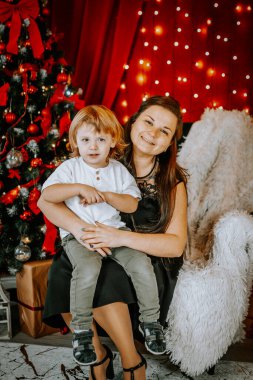 Gülümseyen bir kadın, Noel ağacının yanında, Noel ışıkları ve dekorasyonlarla çevrili bir çocuğu kucağında tutuyor..