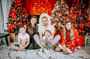 Güzel dekore edilmiş Noel ağaçlarının yanında birlikte poz veren neşeli bir aile, festival ışıkları, süsler ve tatil dekorlarıyla çevrili..