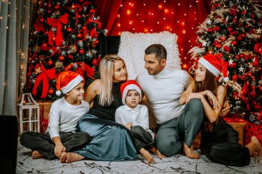 Noel Baba şapkalarında Noel Baba 'nın süslü Noel ağacının yanında toplanan neşeli bir aile. Işıklar, hediyeler ve bayram neşesiyle çevrili. Birlikte poz veriyorlar..