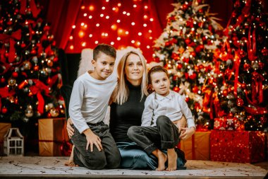 İki oğluyla rahat bir Noel ortamında poz veren bir anne. Etrafı ışıklı ağaçlar, kırmızı süsler ve şenlikli süslemelerle çevrili..