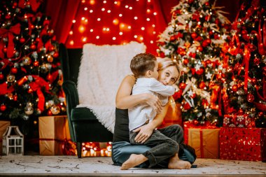 Bir anne, küçük oğlunu sevgi dolu bir Noel süslemesi ile kucaklar. Işıklı ağaçlar, kırmızı süslemeler ve etrafına hediyeler sarar..