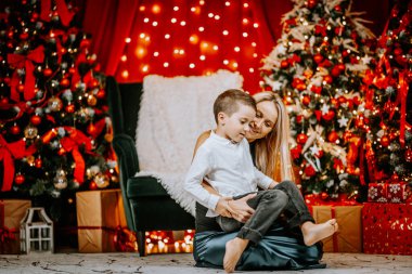 Sevgi dolu bir anne oğlunu sıcak, şenlikli bir Noel ortamında kucaklar. Parlayan ışıklar, dekore edilmiş ağaçlar ve arka planda paketlenmiş hediyelerle..