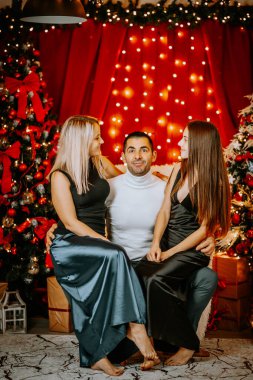 Bir baba, kızıyla karısının arasında ışıldayan ışıklar, süslenmiş ağaçlar ve neşeli ifadelerle şenlikli bir Noel ortamında oturur..